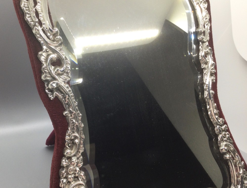 A late Victorian sterling silver easel mirror with embossed floral scrollwork, bevelled glass, and burgundy velvet backing. View X
