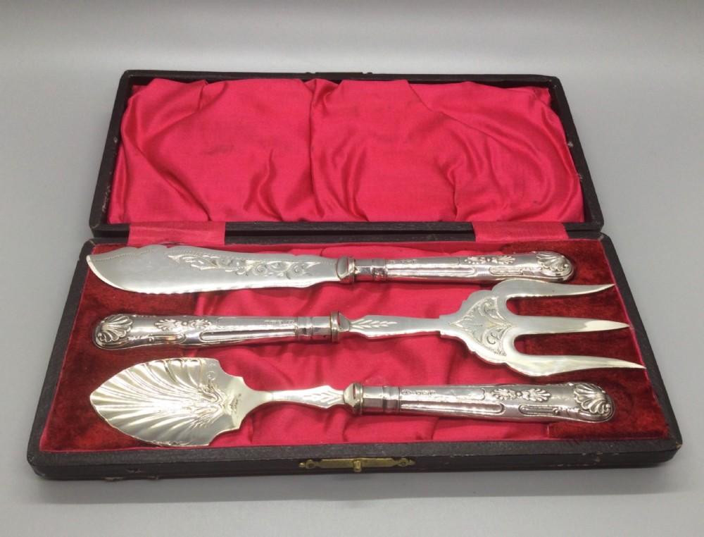 A three-piece Edwardian sterling silver serving set comprising a butter knife, pickle fork, and preserve spoon housed inside an open red velvet-lined presentation case.