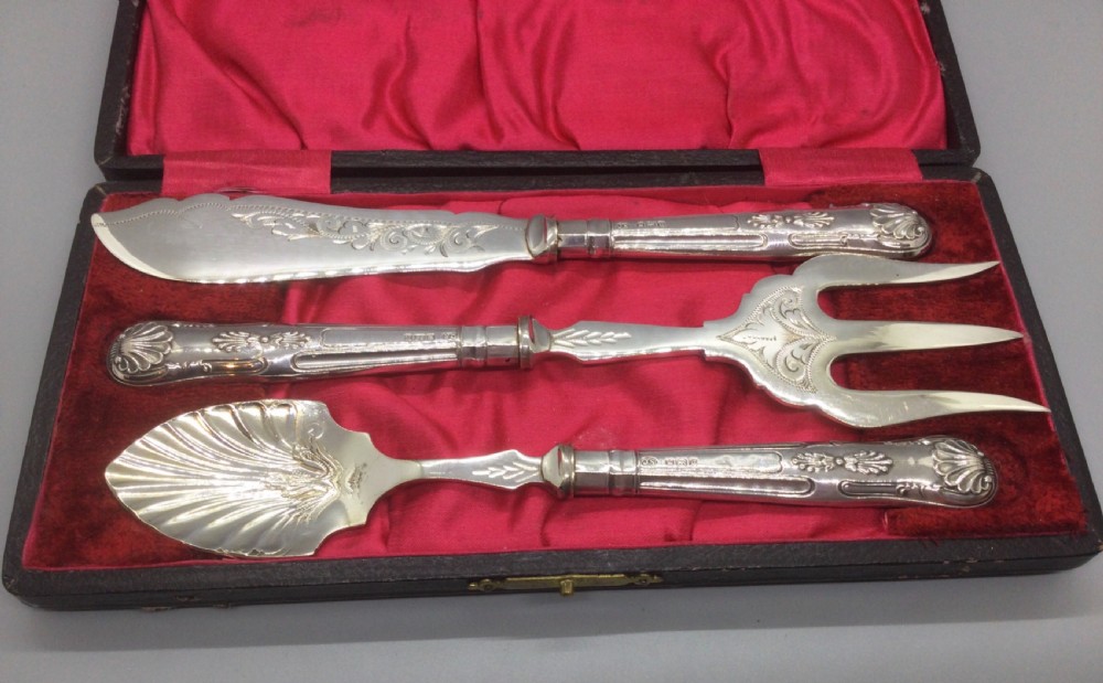 A three-piece Edwardian sterling silver serving set comprising a butter knife, pickle fork, and preserve spoon housed inside an open red velvet-lined presentation case. View II