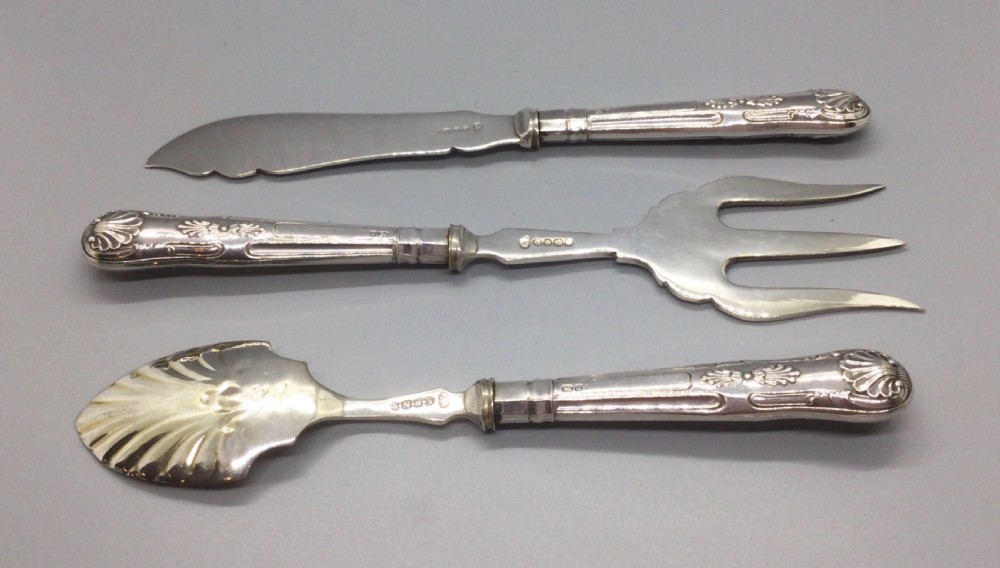 A three-piece Edwardian sterling silver serving set comprising a butter knife, pickle fork, and preserve spoon housed inside an open red velvet-lined presentation case. View VIII