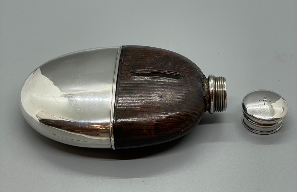 A Victorian spirit flask featuring a glass body encased in dark brown leather with a central sight window and a polished silver lower cup and cap. View X