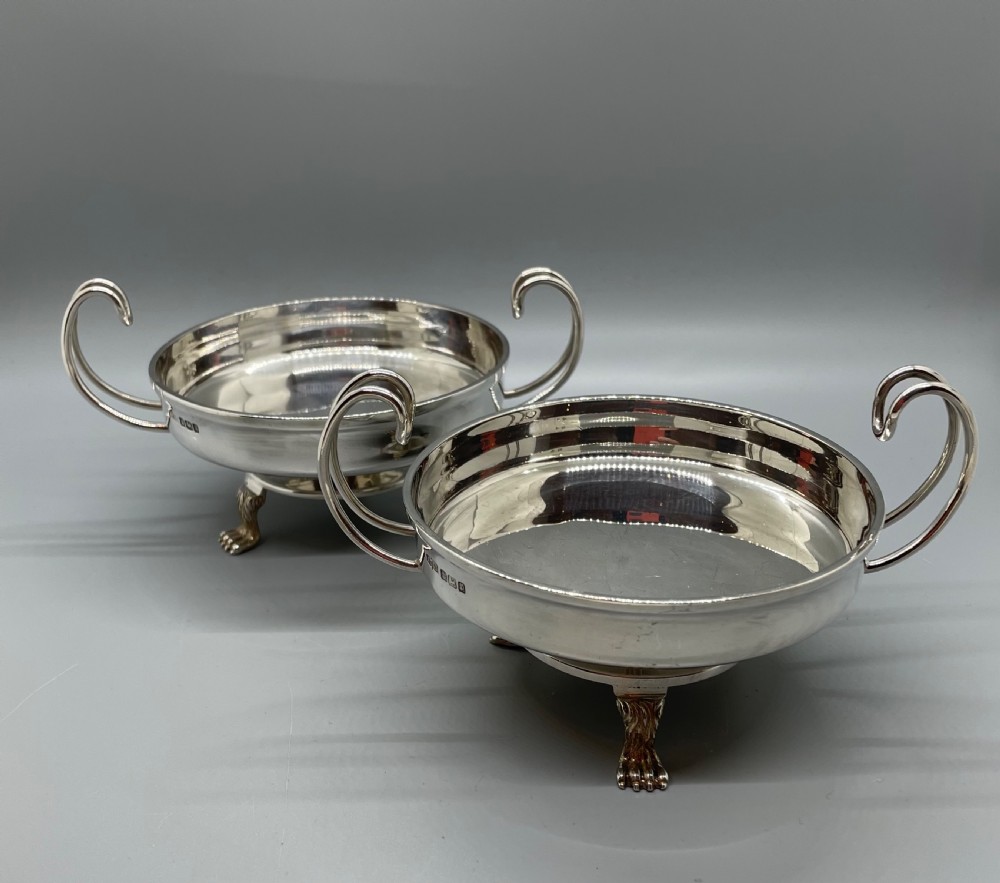 A pair of round Art Deco sterling silver bonbon dishes featuring twin scroll handles, lion paw feet, and visible Sheffield hallmarks. View V