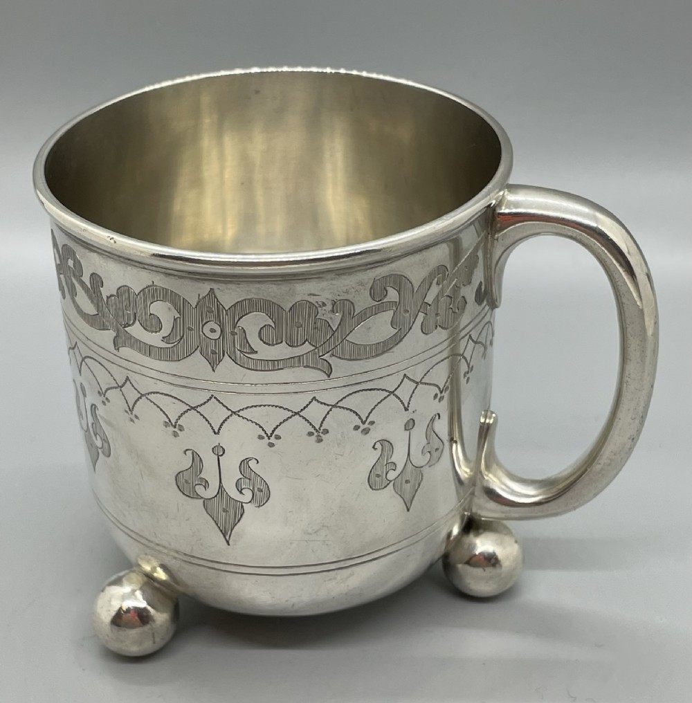 A Victorian sterling silver christening mug featuring hand-engraved scrollwork and geometric patterns, supported by three spherical ball feet.