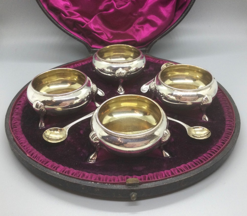 A set of four Victorian sterling silver open salt cellars with gilt interiors and shell-capped feet, displayed in a velvet-lined case with spoons.