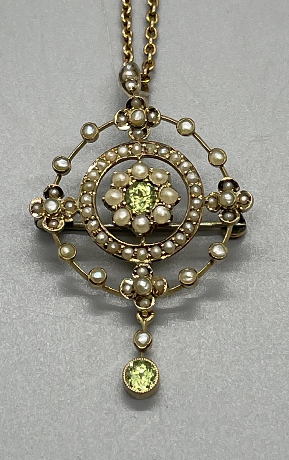 An Edwardian 9ct gold circular openwork pendant set with seed pearls and round faceted peridots, featuring a hanging drop gemstone and attached chain.