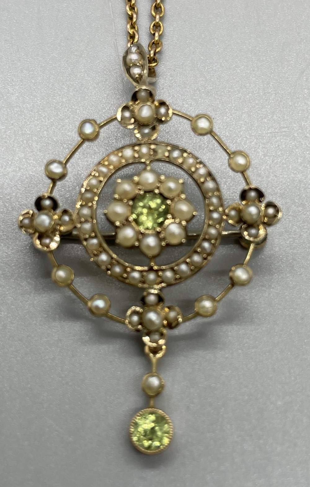 An Edwardian 9ct gold circular openwork pendant set with seed pearls and round faceted peridots, featuring a hanging drop gemstone and attached chain. View IV