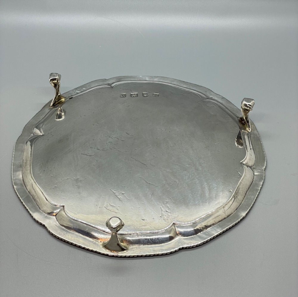 A circular Georgian sterling silver salver featuring a shaped gadrooned rim, an engraved central monogram, and three cast feet. View X