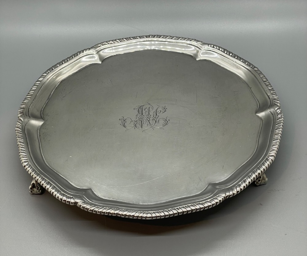 A circular Georgian sterling silver salver featuring a shaped gadrooned rim, an engraved central monogram, and three cast feet. View XI