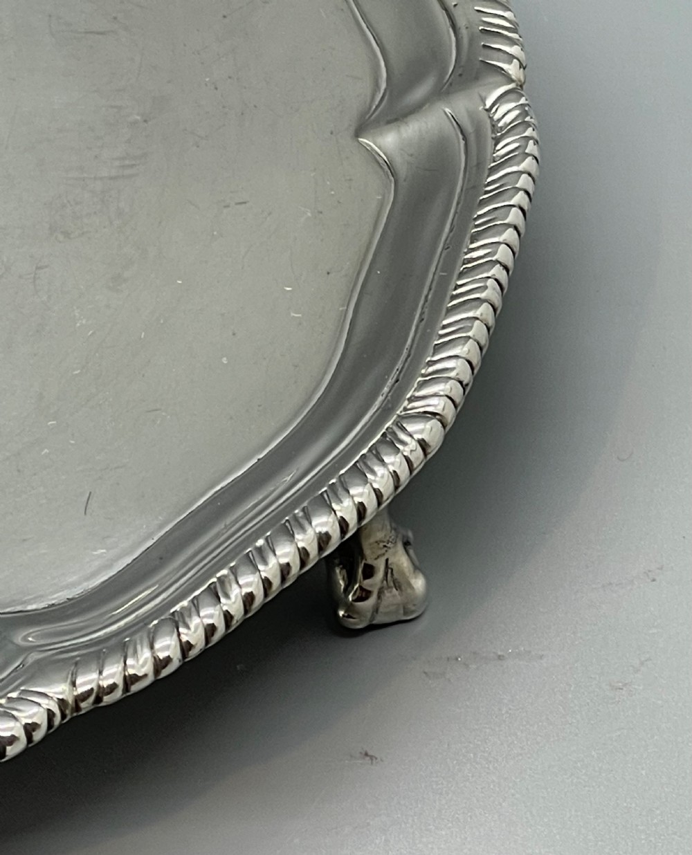 A circular Georgian sterling silver salver featuring a shaped gadrooned rim, an engraved central monogram, and three cast feet. View XV
