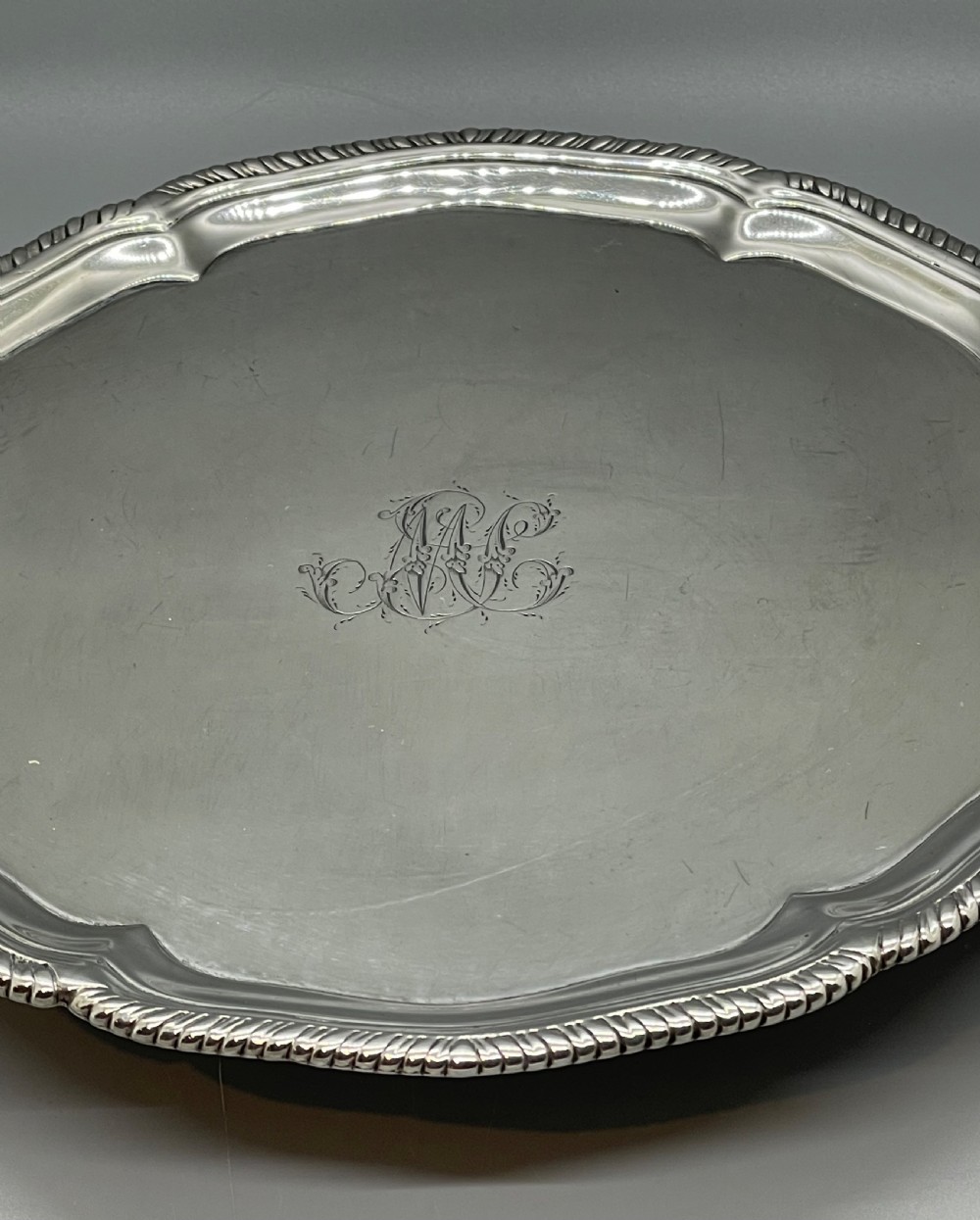 A circular Georgian sterling silver salver featuring a shaped gadrooned rim, an engraved central monogram, and three cast feet. View XVI
