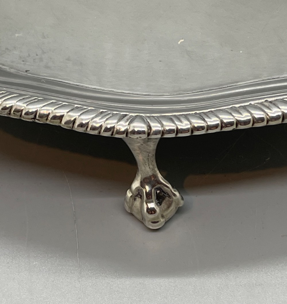 A circular Georgian sterling silver salver featuring a shaped gadrooned rim, an engraved central monogram, and three cast feet. View XVII