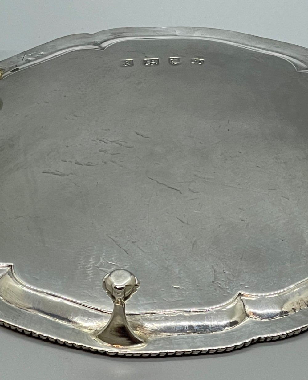 A circular Georgian sterling silver salver featuring a shaped gadrooned rim, an engraved central monogram, and three cast feet. View XXI