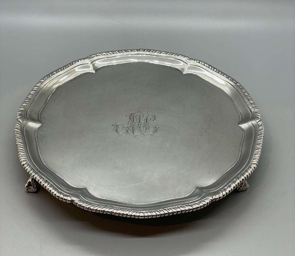 A circular Georgian sterling silver salver featuring a shaped gadrooned rim, an engraved central monogram, and three cast feet. View XXIII