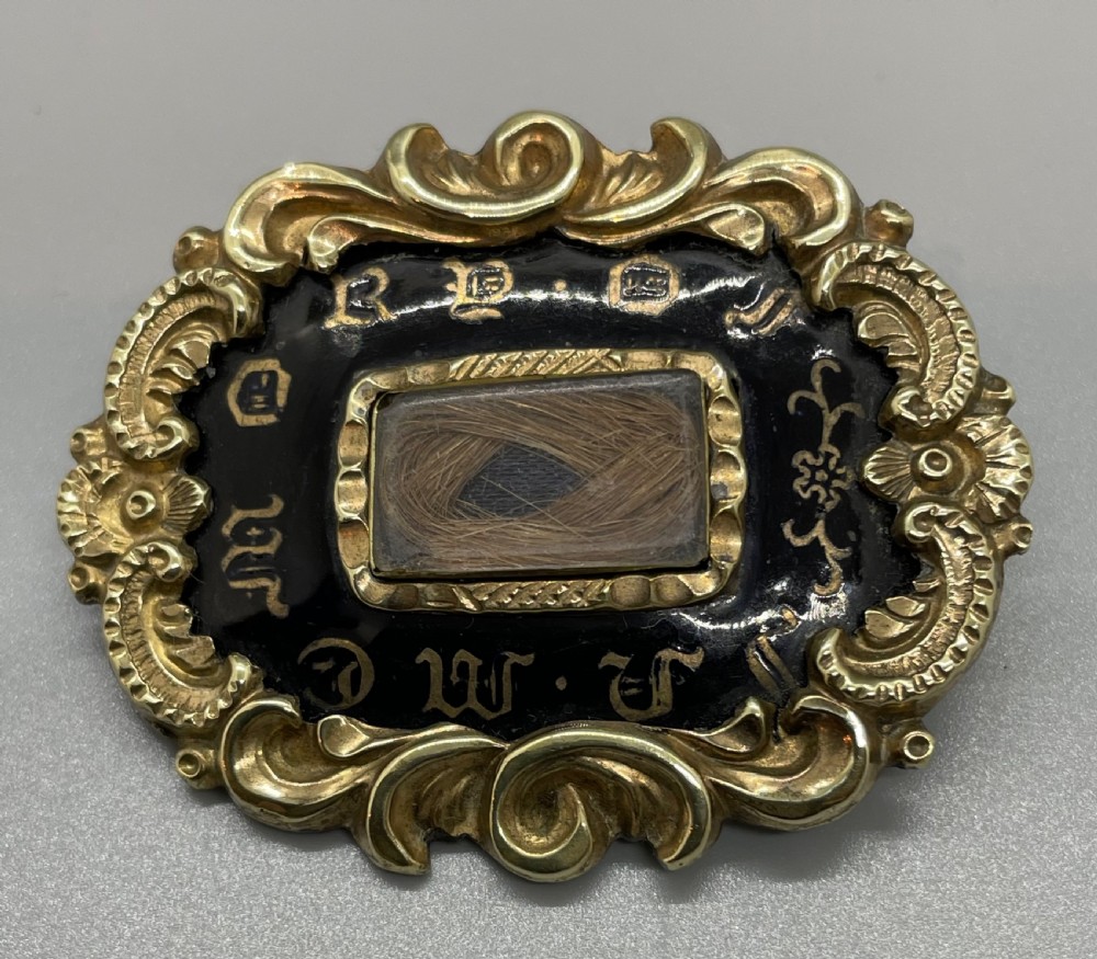 An oval early Victorian gold mourning brooch featuring a scrollwork frame, black enamel border with lettering, and a glass compartment containing woven hair.