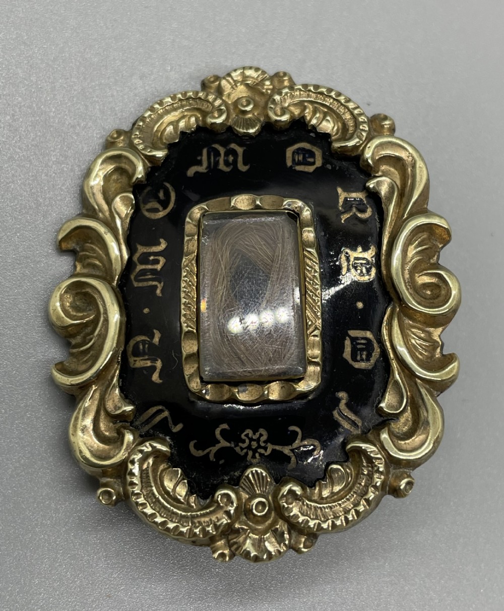 An oval early Victorian gold mourning brooch featuring a scrollwork frame, black enamel border with lettering, and a glass compartment containing woven hair. View II