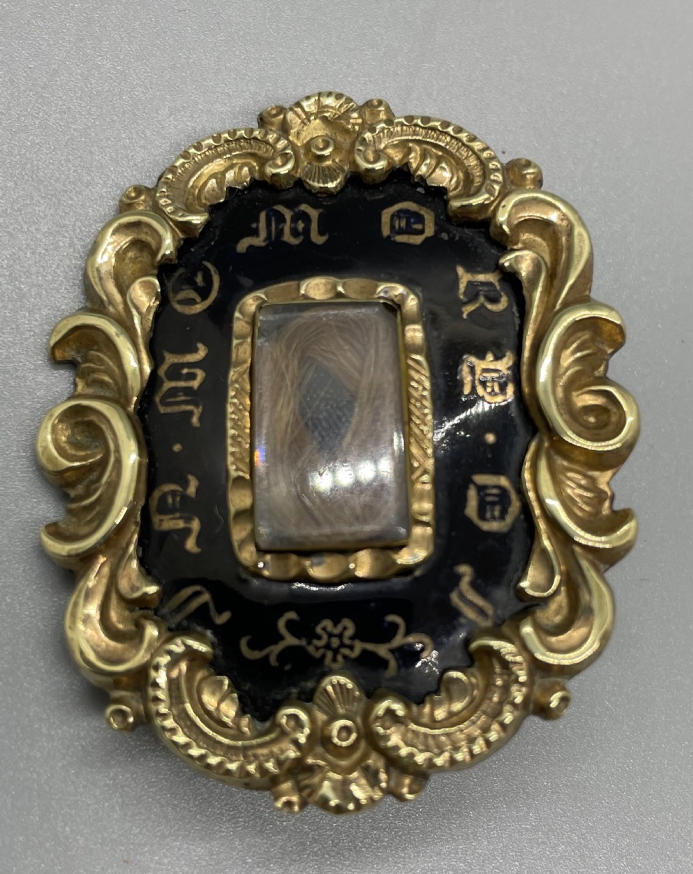 An oval early Victorian gold mourning brooch featuring a scrollwork frame, black enamel border with lettering, and a glass compartment containing woven hair. View III