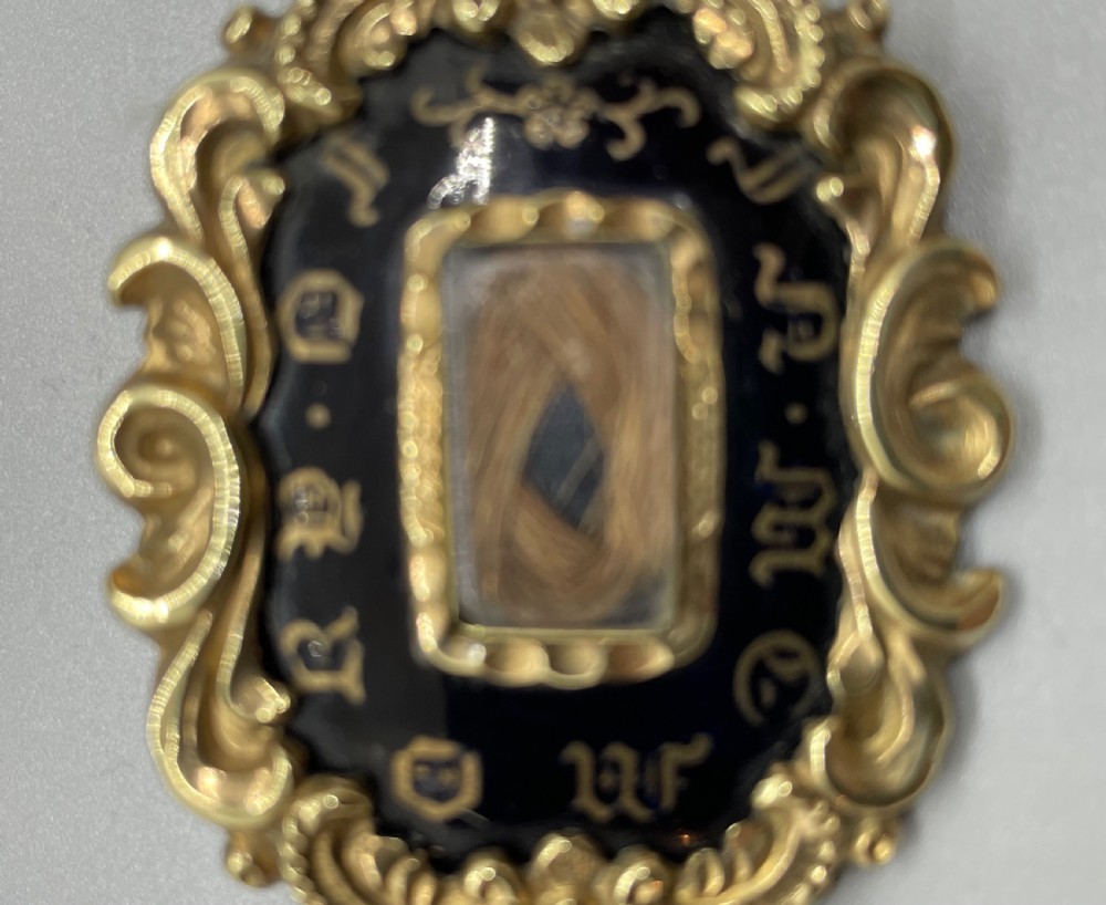 An oval early Victorian gold mourning brooch featuring a scrollwork frame, black enamel border with lettering, and a glass compartment containing woven hair. View V