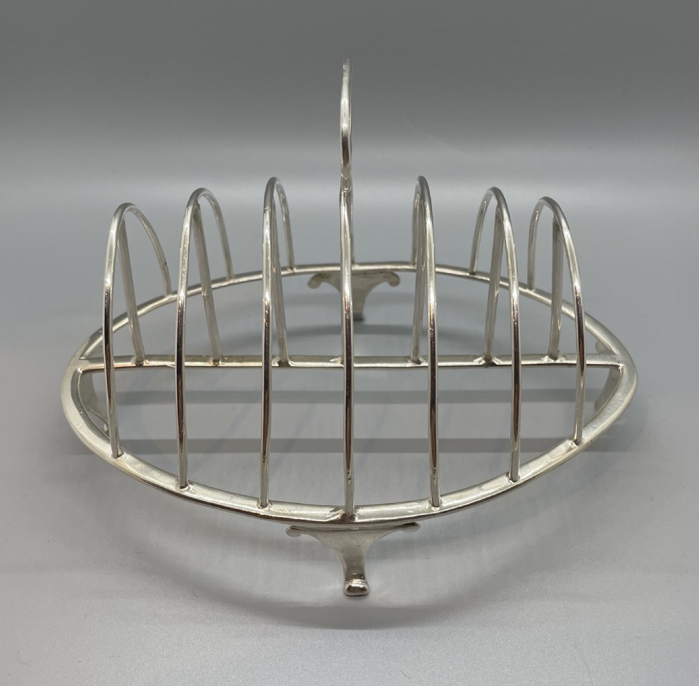 A George III sterling silver toast rack featuring seven arched divisions, an oval base frame, bracket feet, and a central ring handle. View III