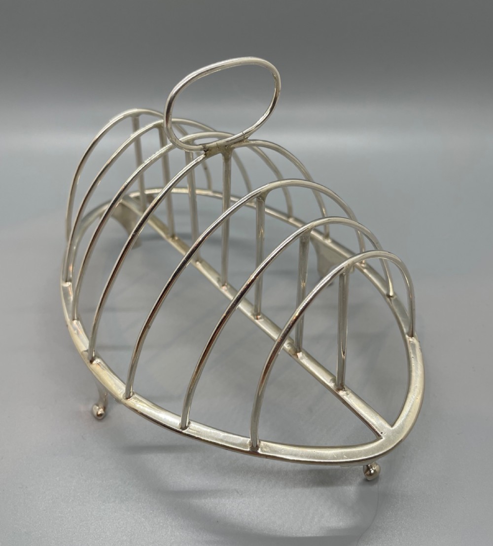 A George III sterling silver toast rack featuring seven arched divisions, an oval base frame, bracket feet, and a central ring handle. View V