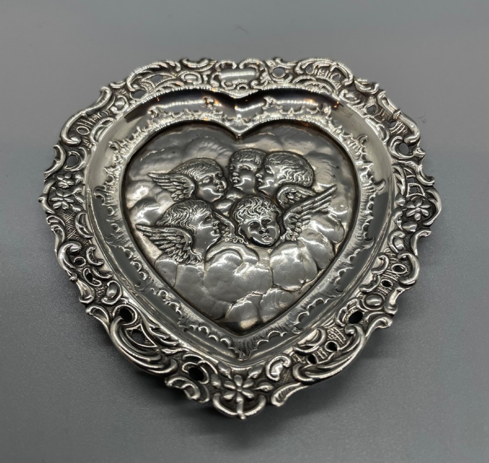 A late Victorian heart-shaped sterling silver trinket dish featuring an embossed central motif of cherub heads amongst clouds with a pierced scroll border.