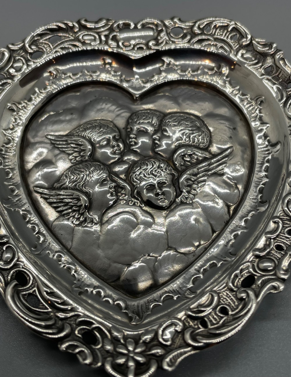 A late Victorian heart-shaped sterling silver trinket dish featuring an embossed central motif of cherub heads amongst clouds with a pierced scroll border. View II