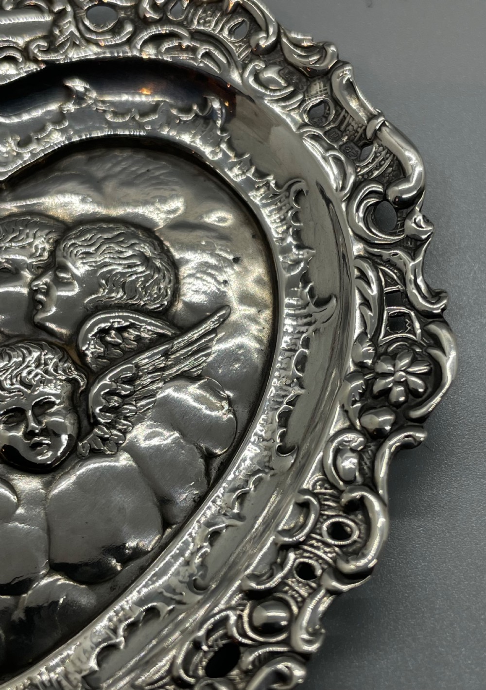 A late Victorian heart-shaped sterling silver trinket dish featuring an embossed central motif of cherub heads amongst clouds with a pierced scroll border. View VI