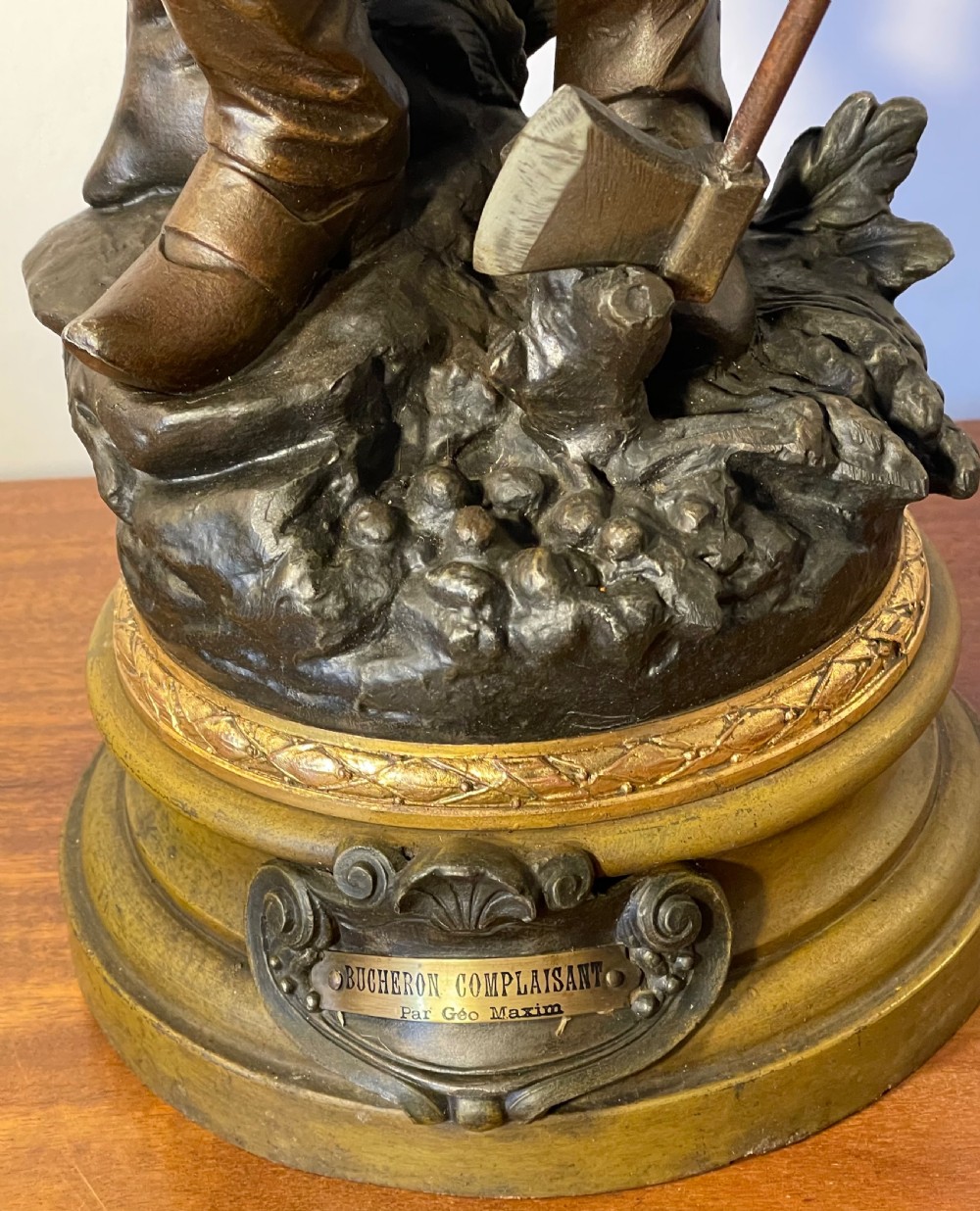 A dark patinated bronze figure depicting a standing woodsman holding an axe on a circular cast base with a named plaque. View VI