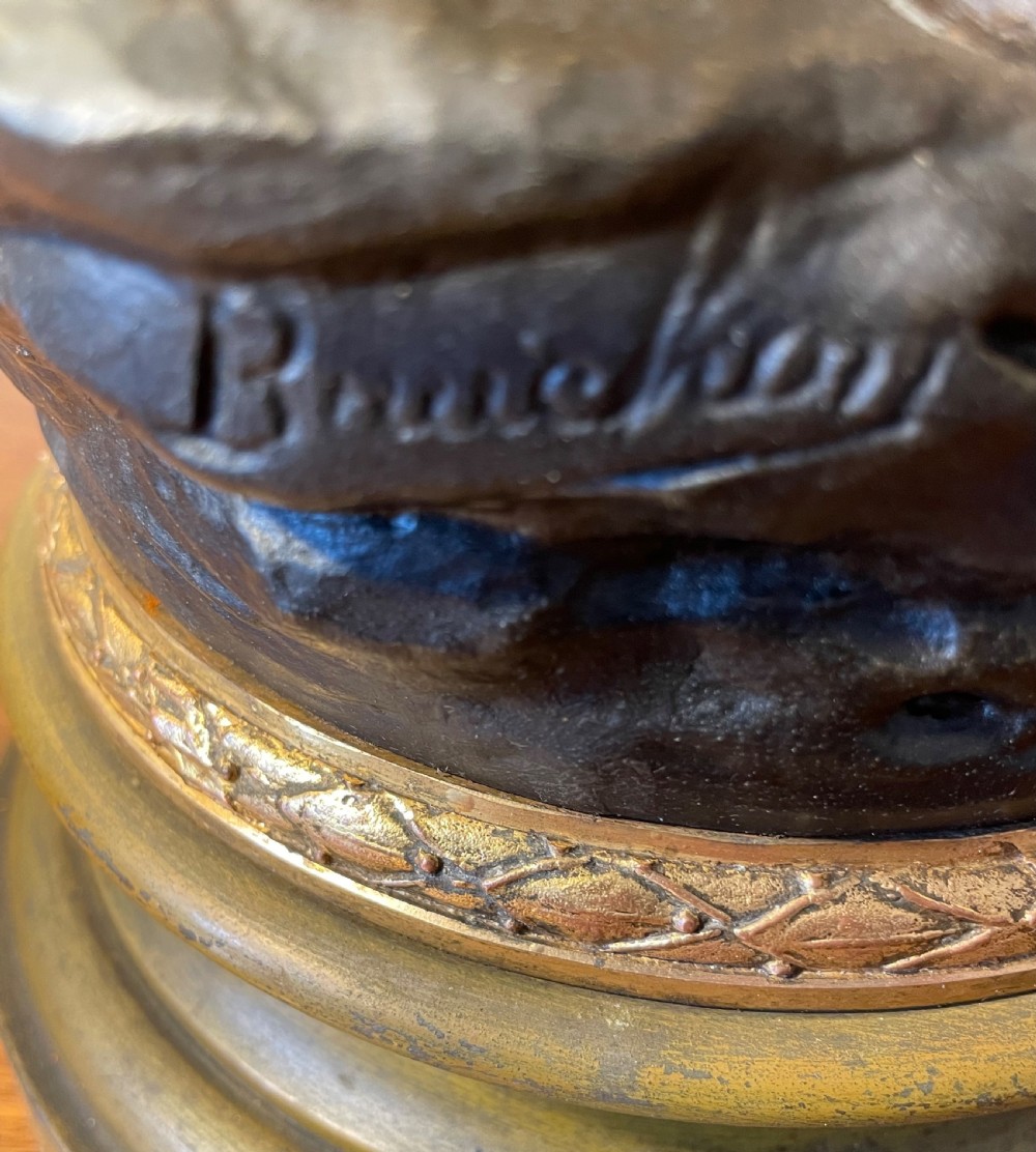A dark patinated bronze figure depicting a standing woodsman holding an axe on a circular cast base with a named plaque. View XII
