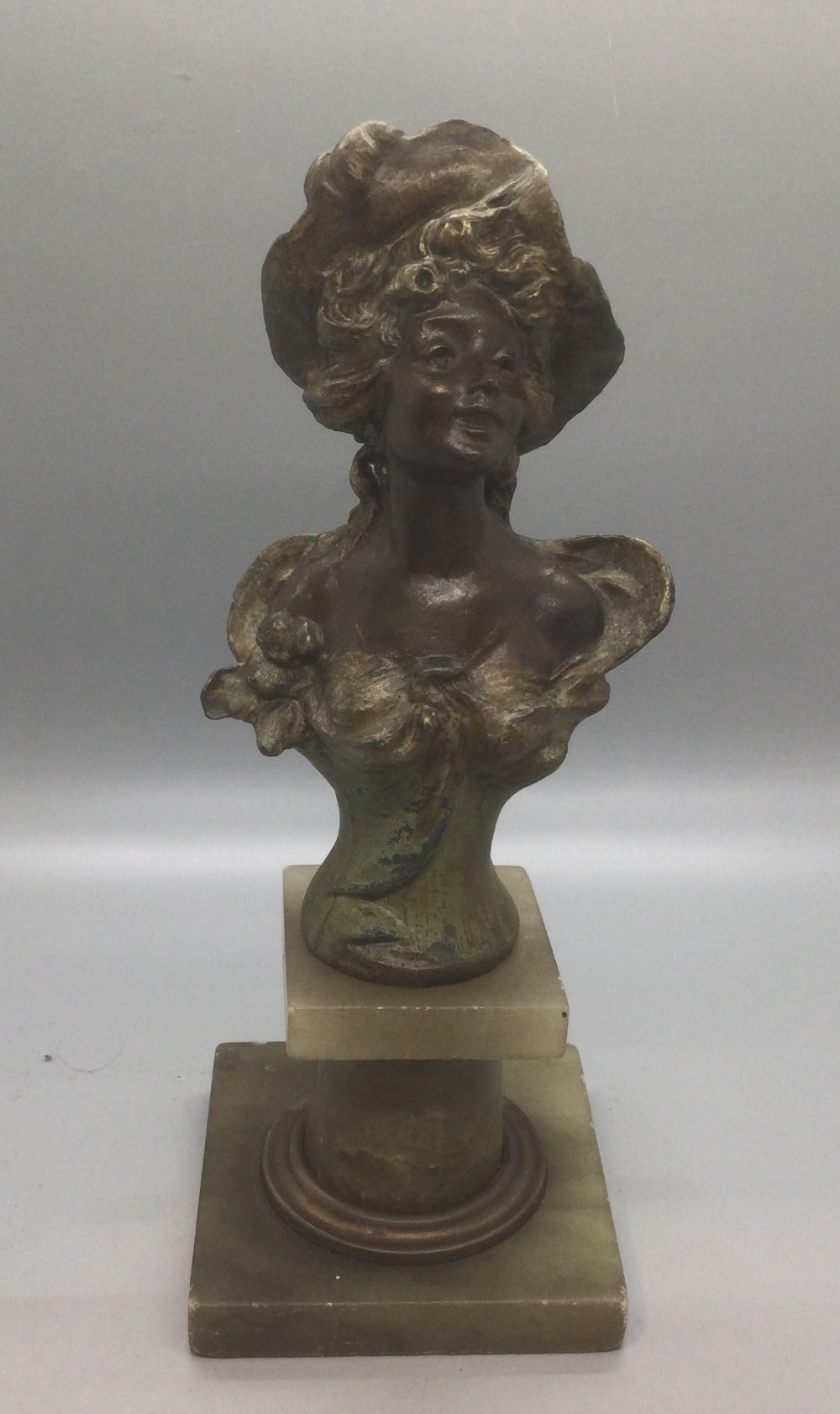 An Art Nouveau patinated spelter bust depicting a female figure wearing a wide hat mounted on a stepped alabaster pedestal base.