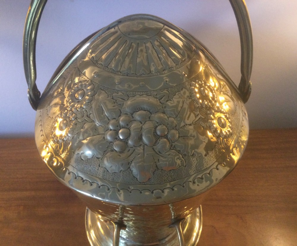 A Victorian brass helmet-shaped coal bucket featuring embossed floral details on the hood, a hammered body, swing handle, and pedestal base. View V