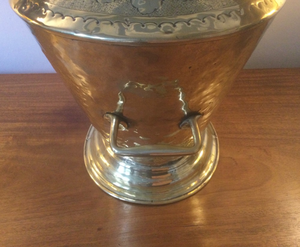 A Victorian brass helmet-shaped coal bucket featuring embossed floral details on the hood, a hammered body, swing handle, and pedestal base. View VI
