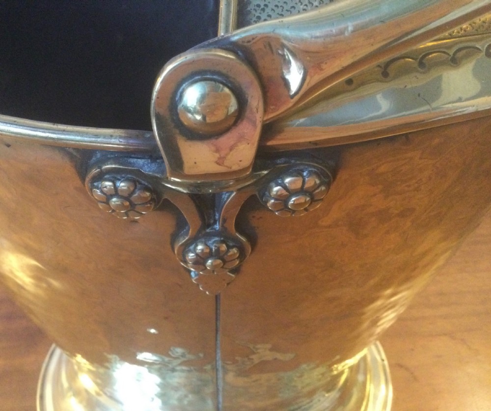 A Victorian brass helmet-shaped coal bucket featuring embossed floral details on the hood, a hammered body, swing handle, and pedestal base. View VII
