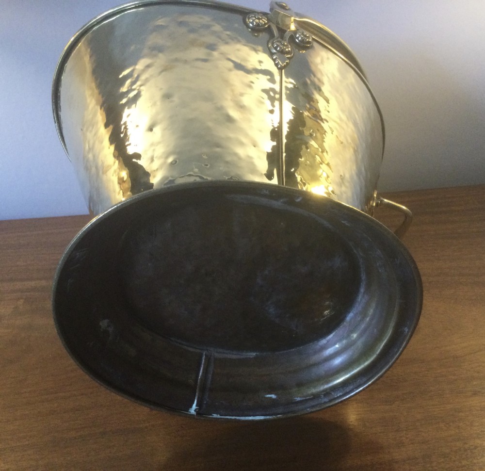 A Victorian brass helmet-shaped coal bucket featuring embossed floral details on the hood, a hammered body, swing handle, and pedestal base. View VIII