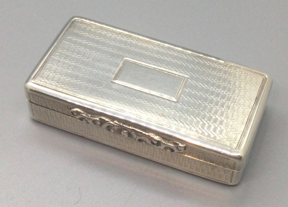 A rectangular Georgian sterling silver snuff box displaying geometric engine-turned engraving, a plain rectangular cartouche, and a chased silver thumbpiece. View II