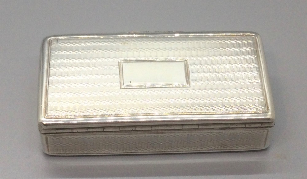 A rectangular Georgian sterling silver snuff box displaying geometric engine-turned engraving, a plain rectangular cartouche, and a chased silver thumbpiece. View IV