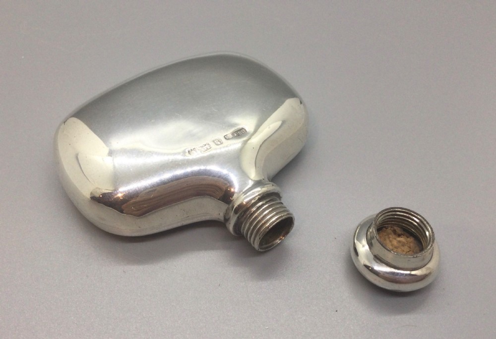 A smooth, polished sterling silver pocket flask with a contoured body and rounded screw cap against a neutral backdrop. View IX