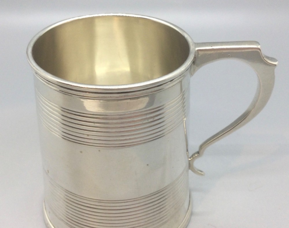 A cylindrical sterling silver christening mug featuring horizontal reeded banding and an angular curved handle. View V