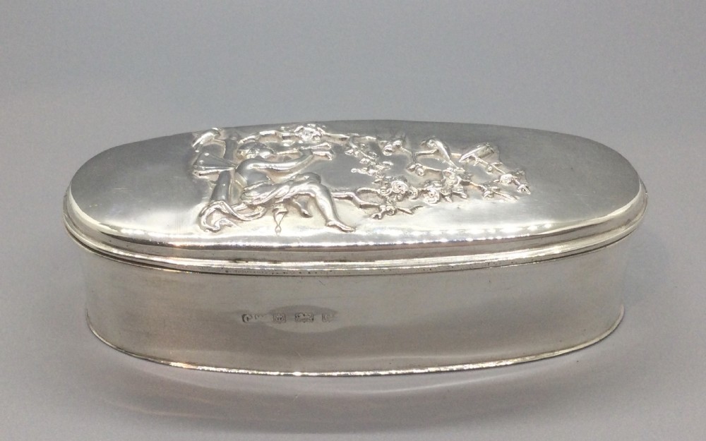 An oval sterling silver box featuring a raised repousse lid depicting a seated figure amongst flowering branches, with visible British hallmarks on the front side.