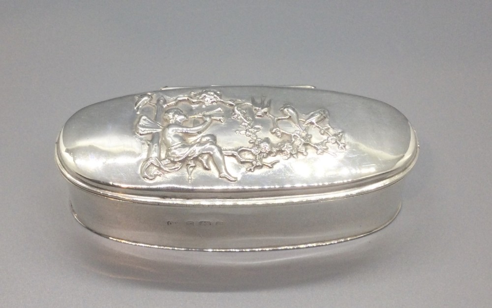 An oval sterling silver box featuring a raised repousse lid depicting a seated figure amongst flowering branches, with visible British hallmarks on the front side. View II
