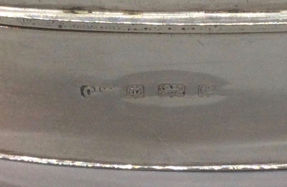 An oval sterling silver box featuring a raised repousse lid depicting a seated figure amongst flowering branches, with visible British hallmarks on the front side. View III