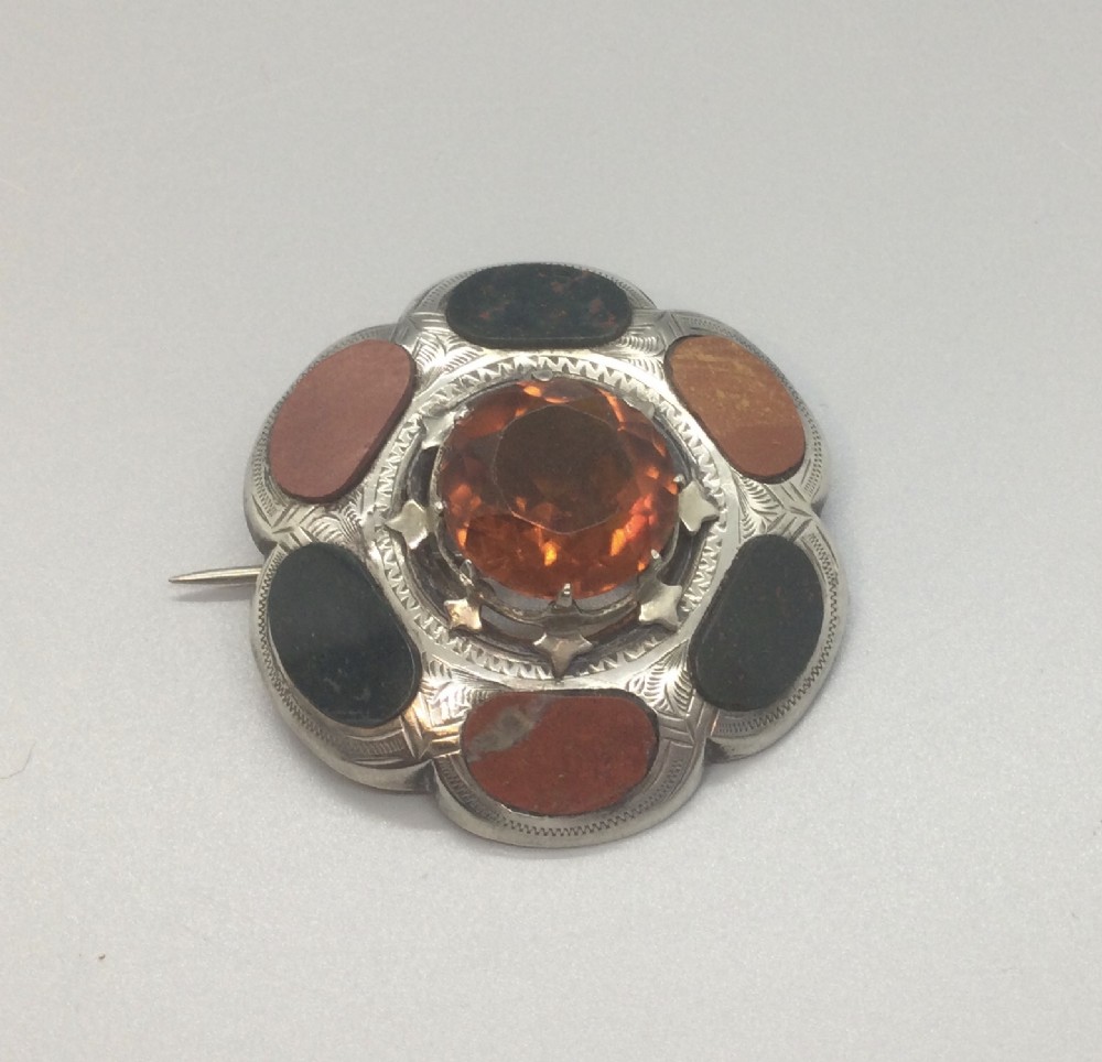 A circular Victorian Scottish silver brooch featuring an engraved scalloped border inlaid with agate stones and set with a central faceted orange citrine.