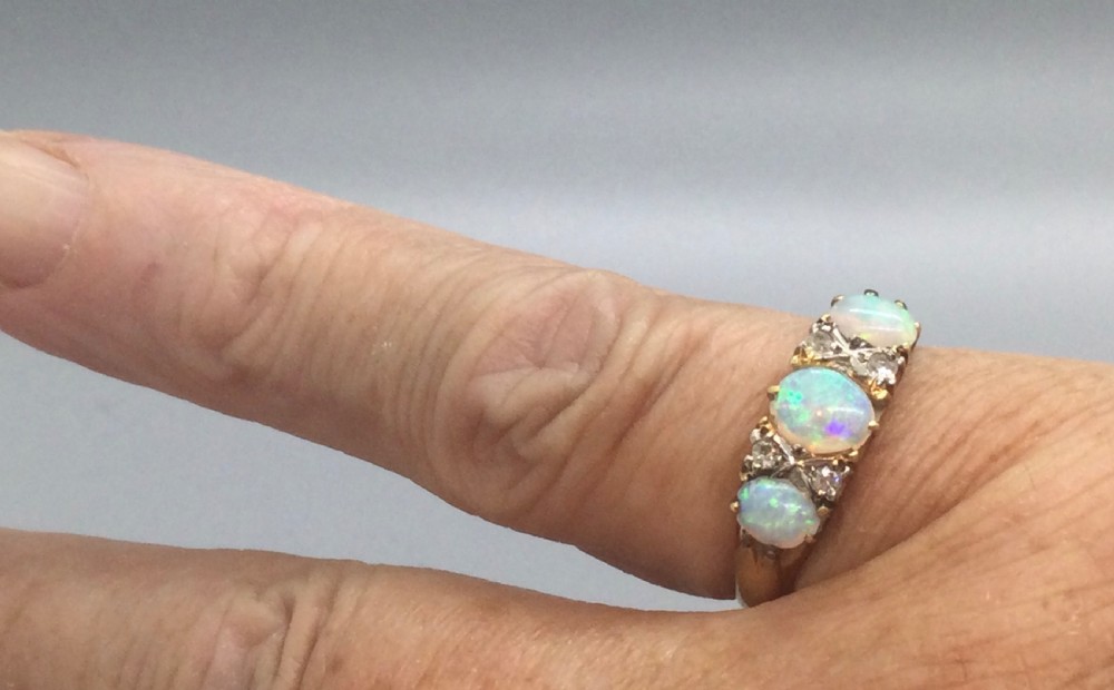 An Edwardian 18ct yellow gold ring featuring three oval cabochon opals separated by diamond accents within a carved gallery. View V