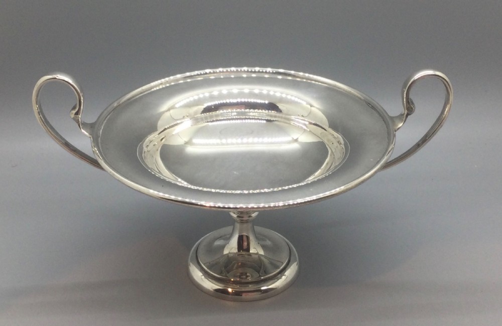 A polished sterling silver pedestal bowl featuring two curved loop handles, a wide shallow dish, and a circular turned foot. View X