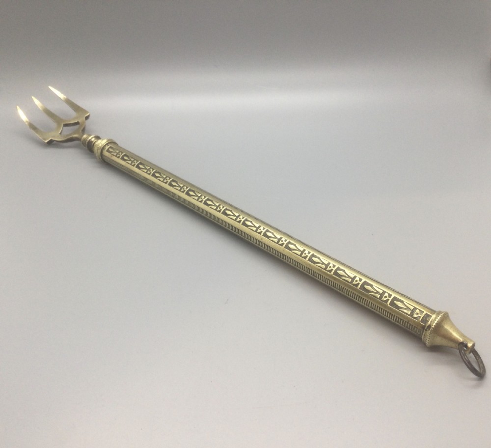 An antique Victorian extendable brass toasting fork with three prongs, an engraved geometric handle, and a hanging loop.