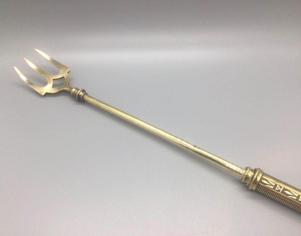 An antique Victorian extendable brass toasting fork with three prongs, an engraved geometric handle, and a hanging loop. View V