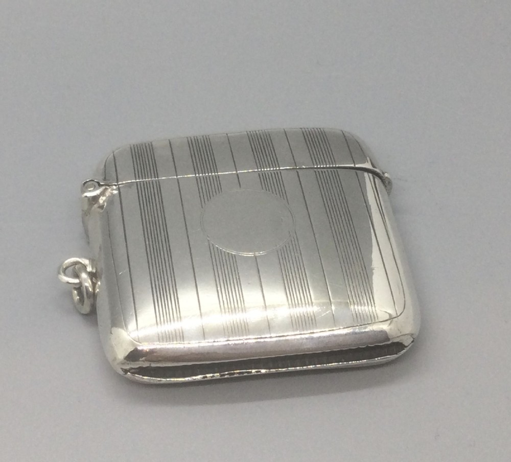 A square sterling silver Art Deco vesta case featuring engine-turned vertical line detailing, a circular central cartouche, a hinged top lid, and a suspension loop on the side.
