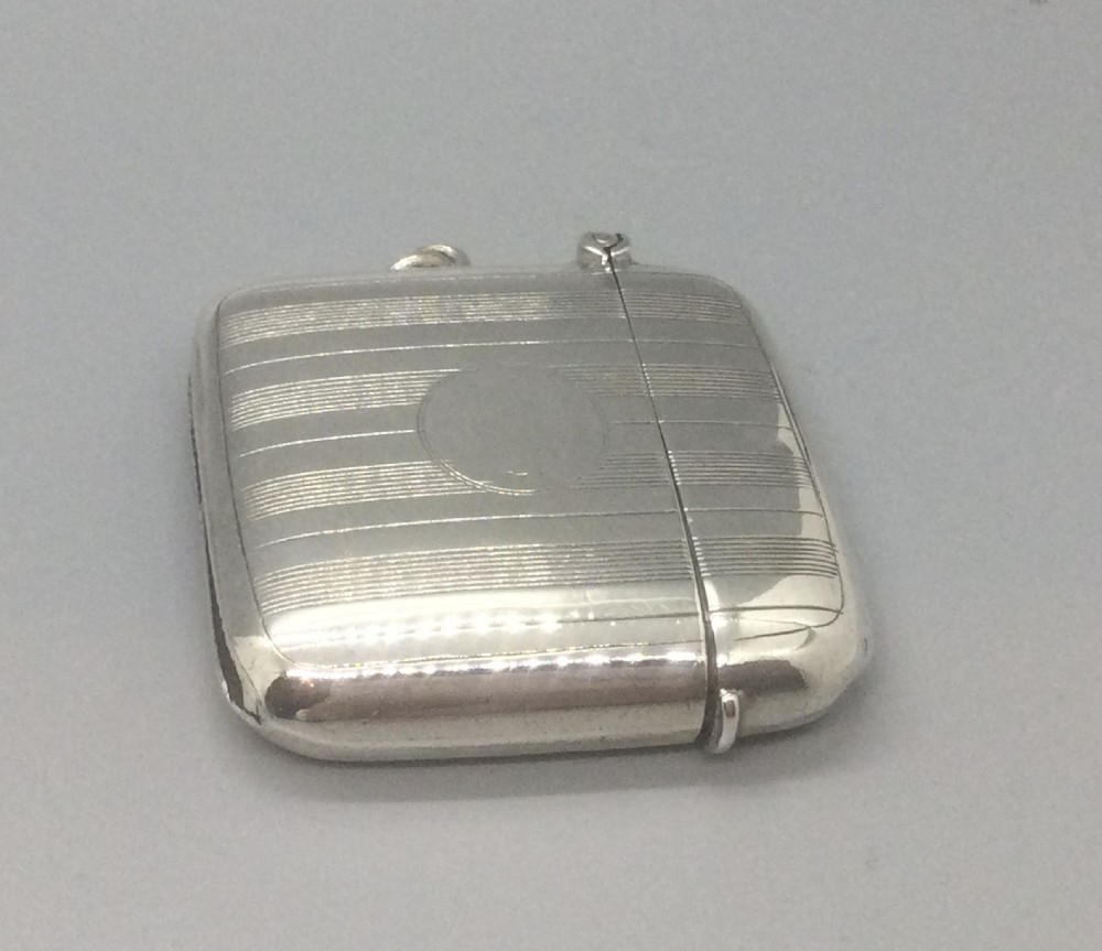 A square sterling silver Art Deco vesta case featuring engine-turned vertical line detailing, a circular central cartouche, a hinged top lid, and a suspension loop on the side. View II
