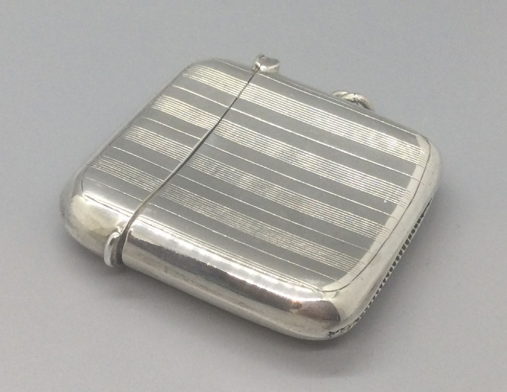 A square sterling silver Art Deco vesta case featuring engine-turned vertical line detailing, a circular central cartouche, a hinged top lid, and a suspension loop on the side. View IV