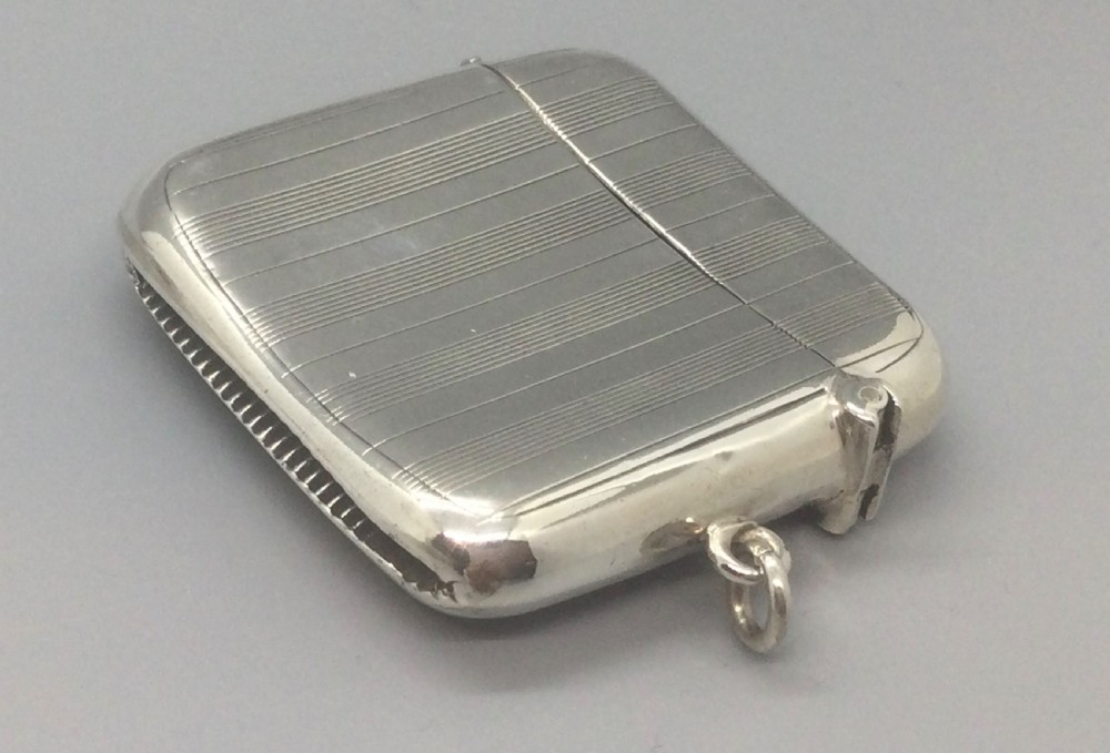 A square sterling silver Art Deco vesta case featuring engine-turned vertical line detailing, a circular central cartouche, a hinged top lid, and a suspension loop on the side. View V