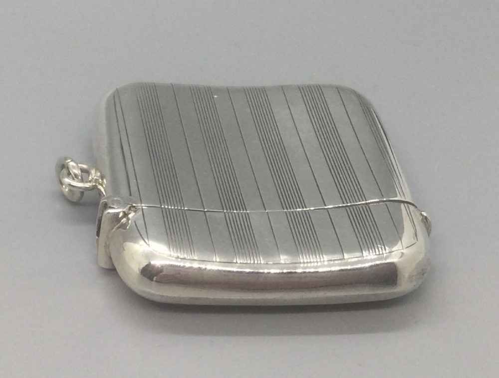 A square sterling silver Art Deco vesta case featuring engine-turned vertical line detailing, a circular central cartouche, a hinged top lid, and a suspension loop on the side. View VI